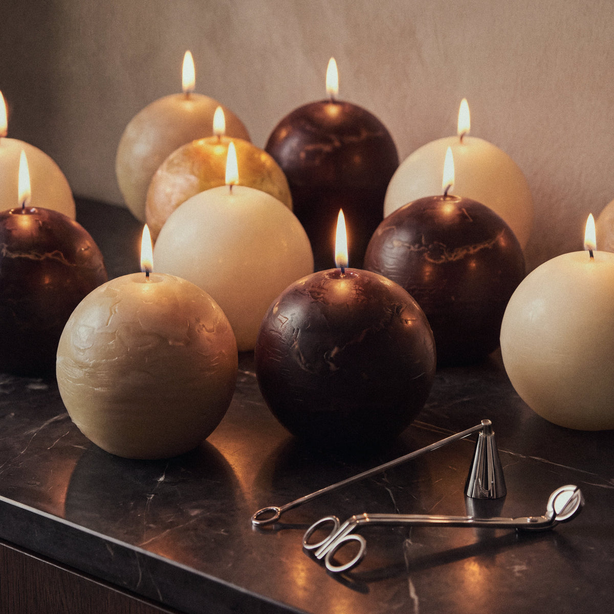 Lit hand-cast sphere candles in neutral shades arranged on a dark marble surface with candle accessories.